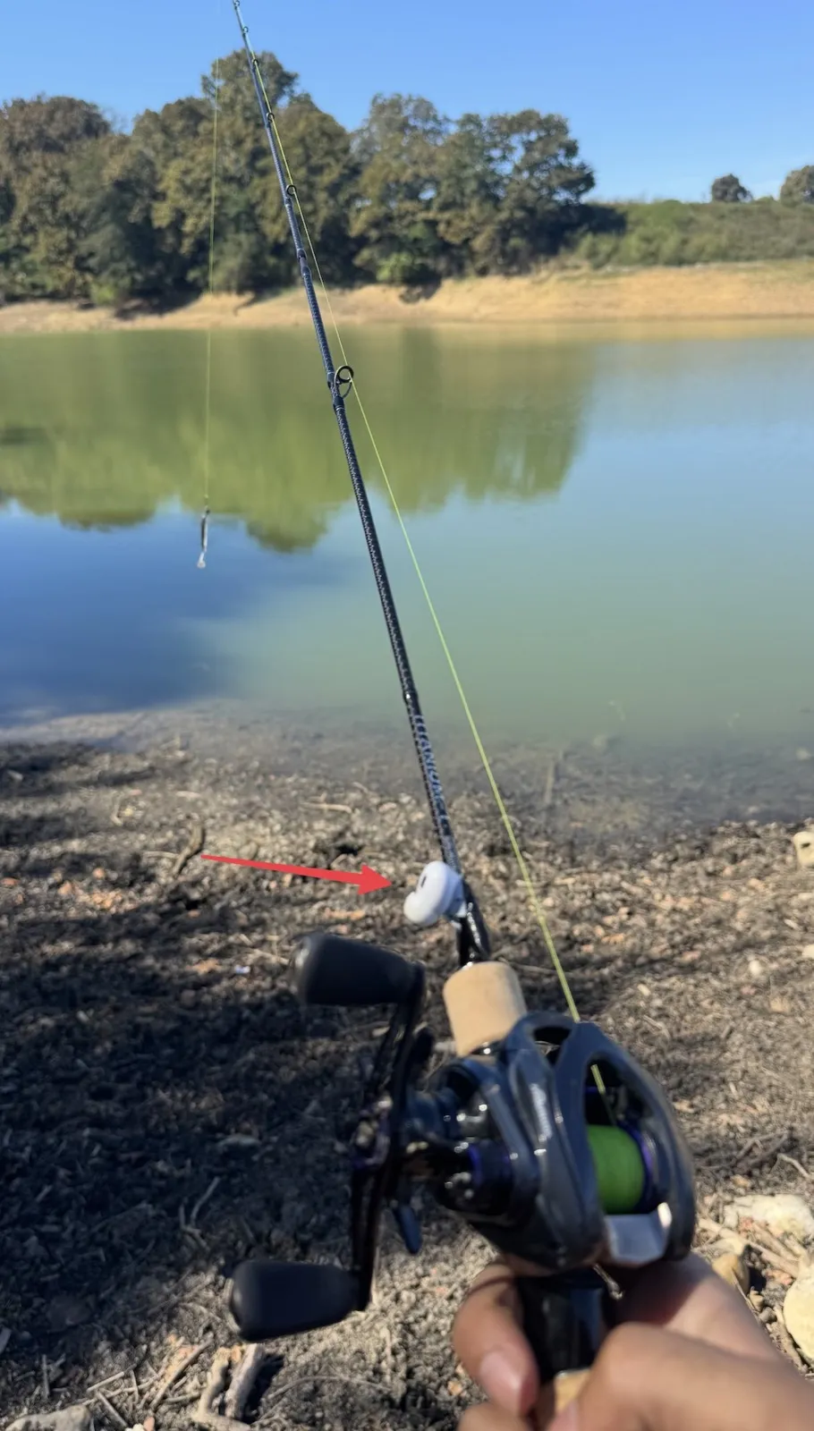 A casting rod held over a lake, with one white AirPod taped to the blank just ahead of the cork grip. A red arrow points to it.